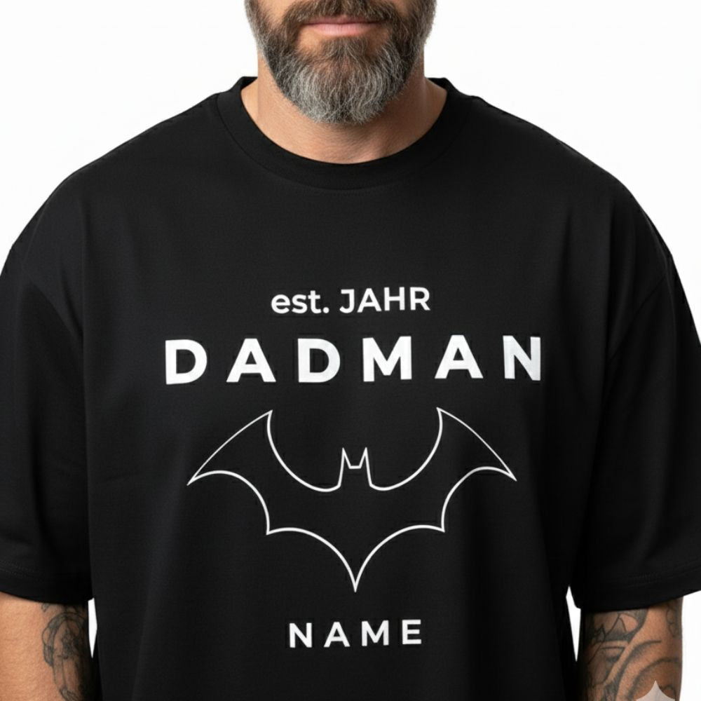 Nahaufnahme des Motivs auf dem PREMIUM Herren T-Shirt "DADMAN" (240 g/m²). Das Design zeigt das personalisierbare Motiv mit "DADMAN", Fledermaus-Logo, "est. JAHR" und "NAME". Detaillierte Ansicht des hochwertigen Drucks auf dem Oversize Shirt.