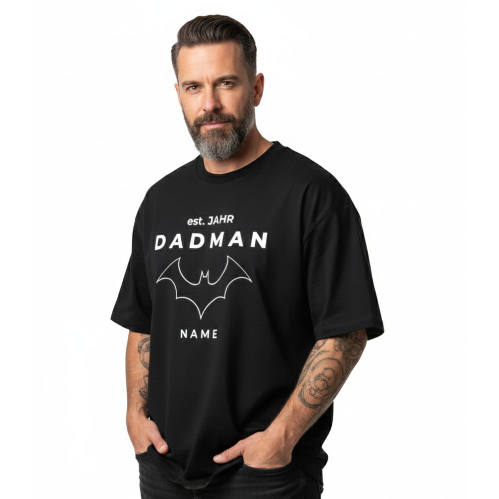 PREMIUM Herren T-Shirt "DADMAN" (240 g/m² Bio-Baumwolle) in Schwarz. Model im lässigen Full-Shot mit Oversize Passform. Papa T-Shirt personalisiert mit "est. JAHR" und "NAME". Ideales Vatertagsgeschenk für stilvolle Väter.