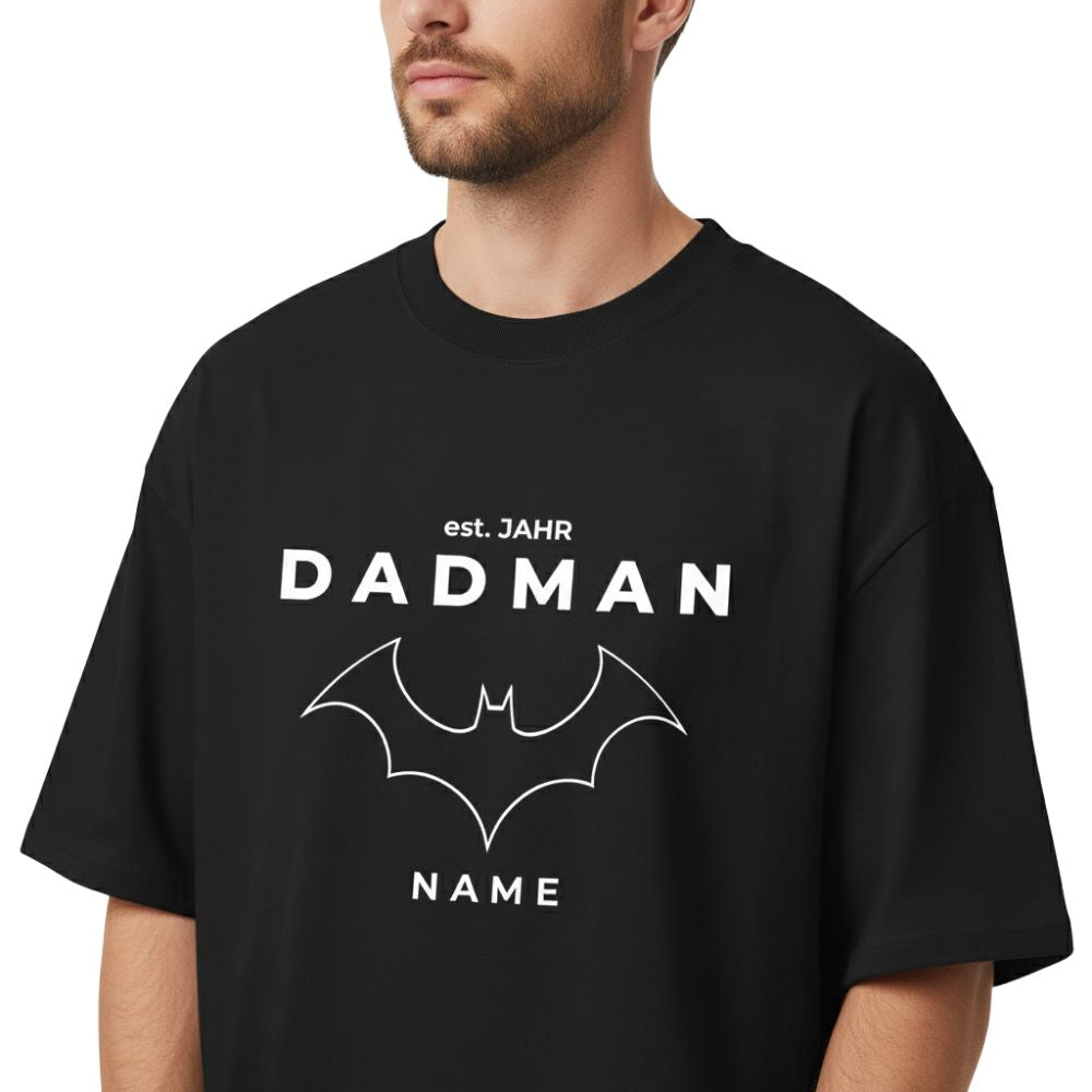 Detailaufnahme (Close-Up) des personalisierten Oversize T-Shirts "DADMAN" in Schwarz. Fokus auf den hochwertigen Druck, den feinen 1x1 Rippstrick-Kragen und den Stoff (200 GSM Bio-Baumwolle). Zeigt die Qualität der Verarbeitung und des Designs.